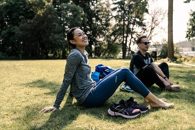 Two people sitting barefoot on the grass in a park after a workout, running shoes off to one side