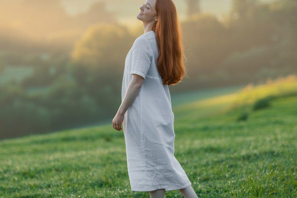 Woman in a white dress walking barefoot through grass at sunrise with her face turned to the light