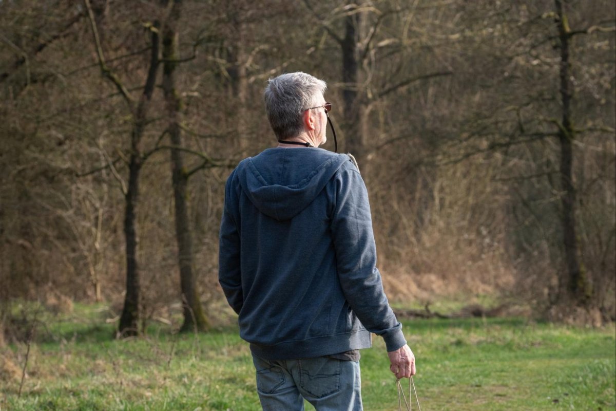 An older man with grey hair walking away across grass toward bare woodland in early spring