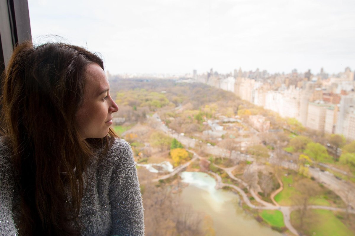 A woman sitting at a high apartment window looking out over a city park below