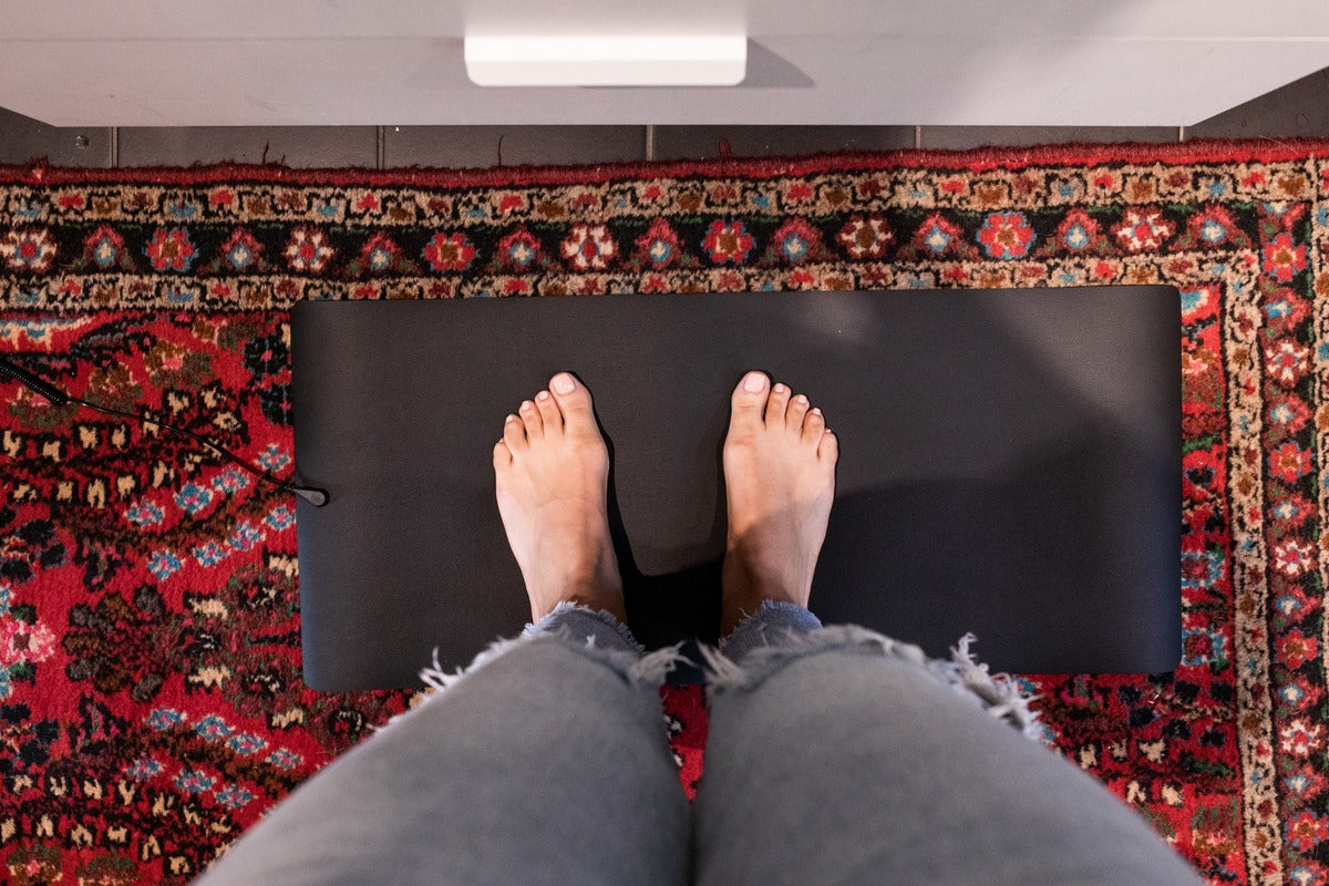 Overhead view of bare feet resting on a black Earthing grounding mat over a patterned rug