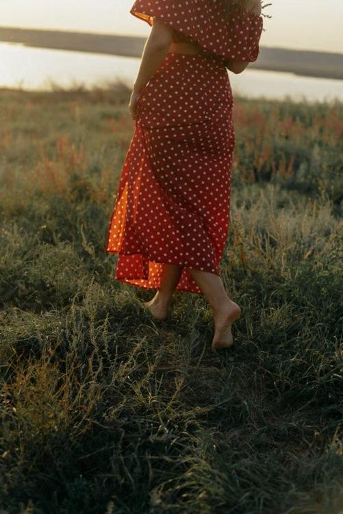 A woman in a red dress walking barefoot through long grass at sunset