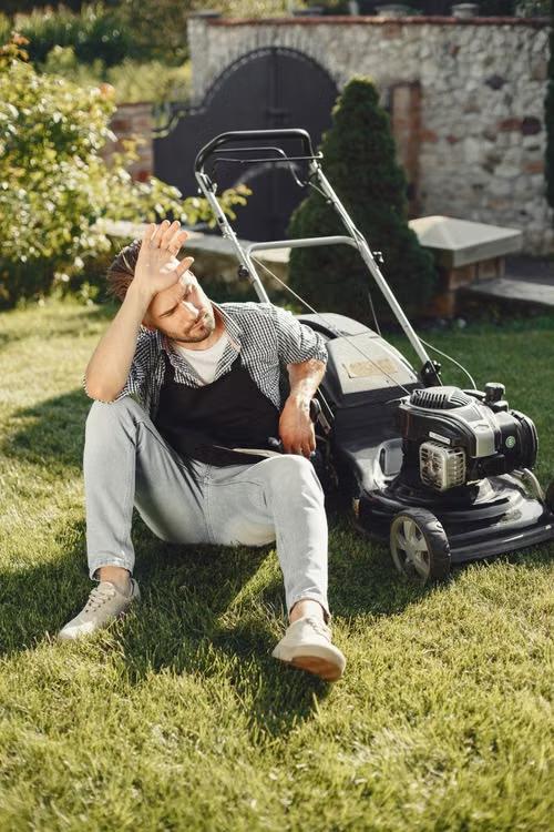 A man sitting on the grass beside a lawn mower, wiping his forehead after yard work