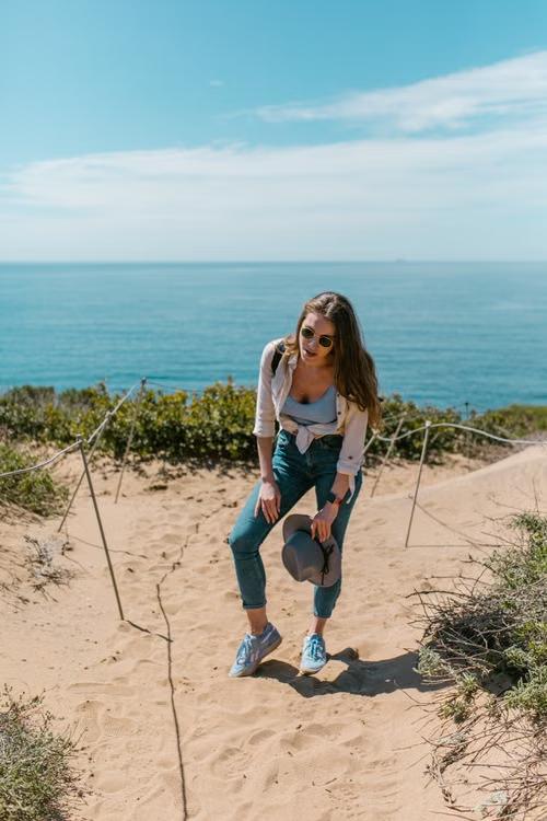 A woman standing barefoot on a sandy path above the sea, holding her hat