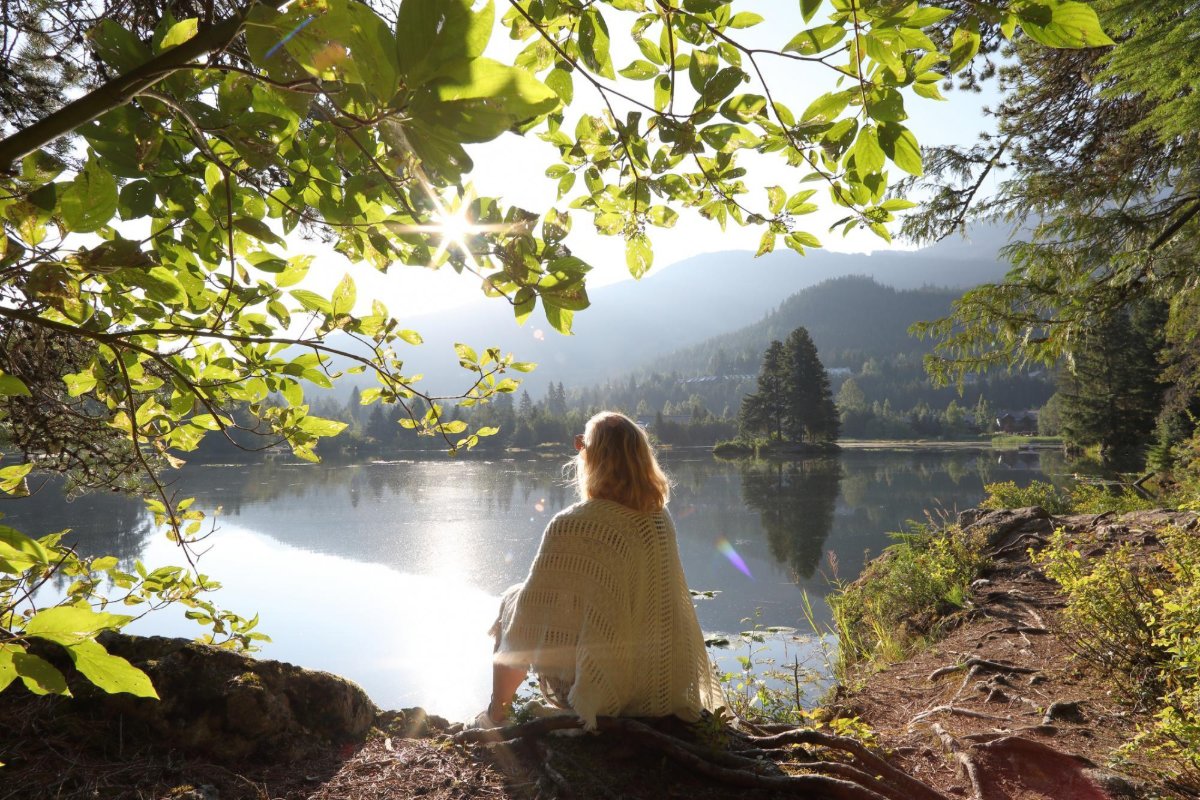 A woman sitting on a rock beside a still mountain lake, framed by overhanging leaves