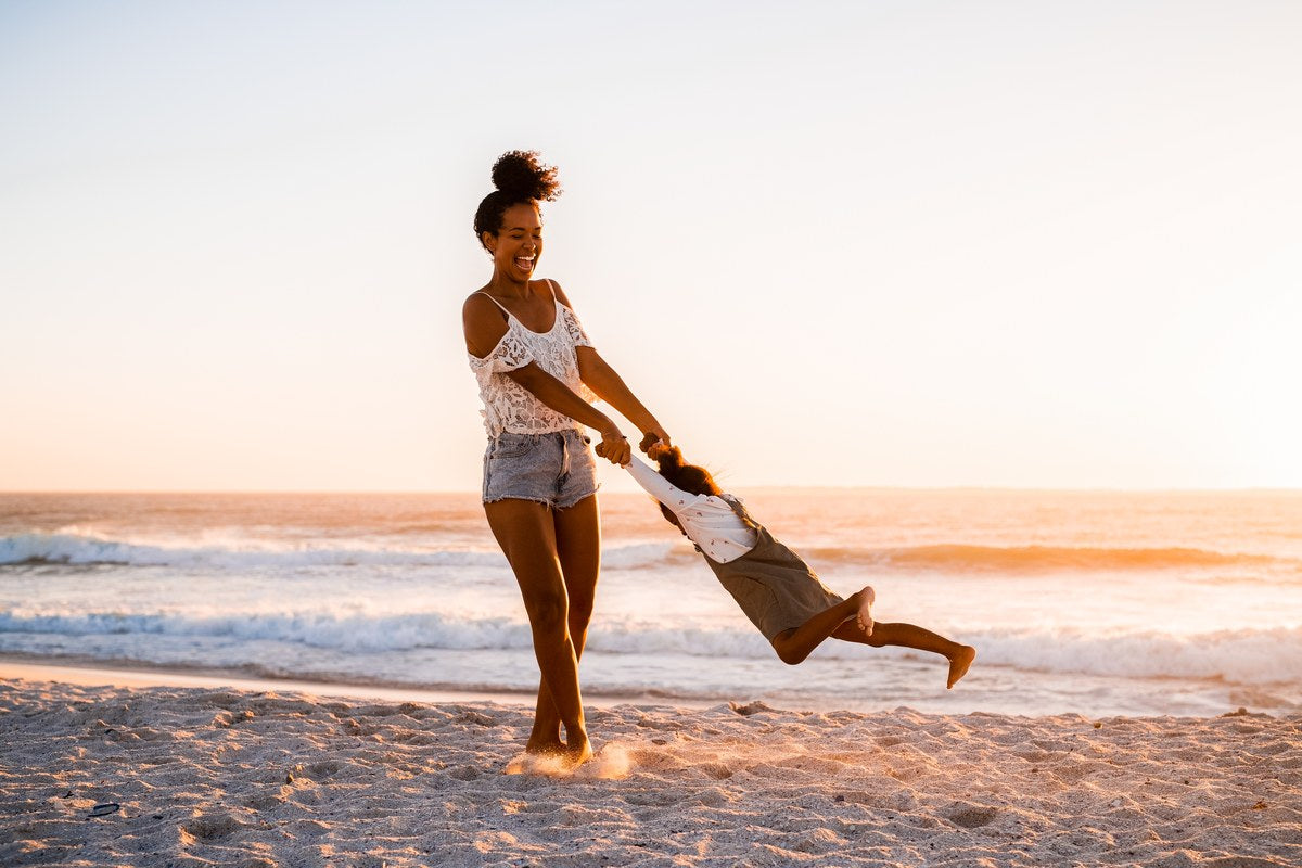 A mother swinging her child barefoot on the sand at sunset