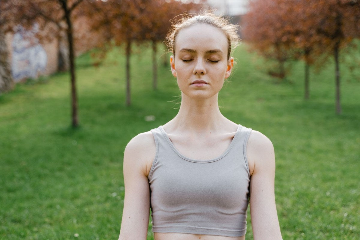 A young woman standing outdoors in a green park with her eyes closed, calm and at rest